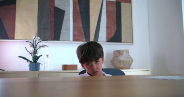 Genç Çocuk üzgün ve kızgın bir ifadeyle masada oturuyor, sakin bir ev ortamında hayal kırıklığı yaşıyor, aile ortamında duygular ve gerilim hissediyor.