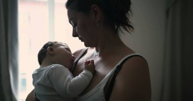 Anne pencerenin kenarında duruyor, bebeği kucaklıyor ve sevgi ve sevgiyle aşağı bakıyor, evde huzurlu ve samimi bir aile anı yakalıyor.