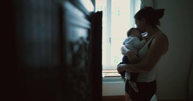 Anne pencerenin kenarında duruyor, bebeği kucaklıyor ve sevgi ve sevgiyle aşağı bakıyor, evde huzurlu ve samimi bir aile anı yakalıyor.