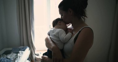 Anne pencerenin kenarında duruyor, bebeği kucaklıyor ve sevgi ve sevgiyle aşağı bakıyor, evde huzurlu ve samimi bir aile anı yakalıyor.