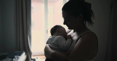 Anne pencerenin yanında dururken bebeğin kucağına usulca sarılıyor. Bağlantının ve aşkın samimi ve huzurlu anını paylaşıyor.