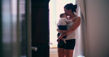 Anne bebeğini hafifçe aydınlatılmış bir odada kucağında tutuyor, şefkatli bir ilgi ve bağlantı anı sergiliyor, sakin bir ortamda sevgiyi ve bakımı ön plana çıkarıyor.