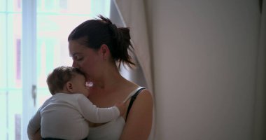 Anne bebeğini kucağında tutuyor, bebeği huzurlu bir ortamda nazikçe kucaklıyor ve rahatlatıyor, sıcaklığı ve bağlantıyı vurguluyor.