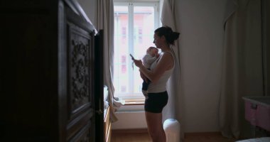 Anne bebeğini akıllı telefon kullanırken kucağında tutuyor, penceresinden gelen yumuşak doğal ışıkla ev ortamında birden fazla iş yapıyor.