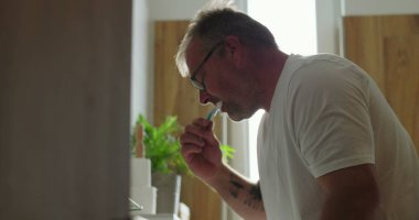 Banyo lavabosunda dişlerini fırçalayan adam, yoğun ifadeli yan profil görüntüsü, parlak ve temiz ev ortamında sabah diş bakımı rutini