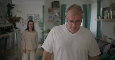 Erkek, arka plandaki kadınla tartışırken gözle görülür şekilde üzgün, oturma odasında ilişki gerilimi, duygusal ve dramatik anlaşmazlık anı