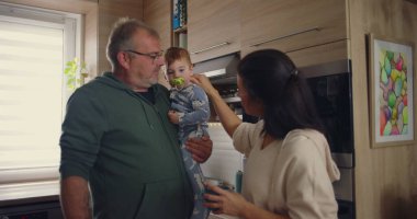 Anne, modern mutfakta babayla konuşurken bebeğin kafasına şefkatle dokunuyor. Günlük ev ortamında sıcak aile etkileşimi.