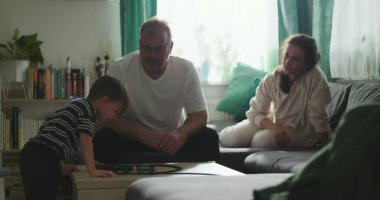 Babam, kahve masasında oyuncak trenle oynayan bir çocukla oturuyor. Anne, kanepeden gülümsüyor ve gözlemliyor. Sıcak aile bağları ve huzurlu ev ortamında samimi bir bağ.