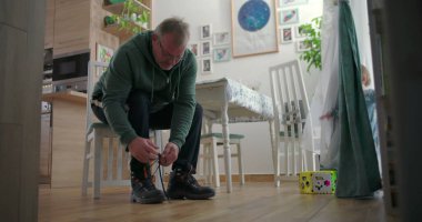 Babam, sıcak ahşap döşemeleri ve doğal ışıklandırması olan sıcak bir aile yemek odasında, emzik ile perdelerin yanında duran bir çocuğa bakarken botlarını bağlıyor.