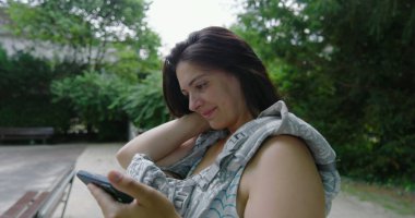 Bebek arabasının önünde bebeğiyle rahat bir anne parkta akıllı telefonunu kontrol ediyor, doğal ortamda sakin bir anın tadını çıkarıyor, teknolojiyi harmanlıyor ve ebeveynlik yapıyor.