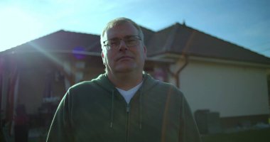 Evin arka bahçesinde duran adam, sakin bir ifade, banliyö manzarası, arka planda yeşil çimenler ve veranda, sakin atmosfer, rahat ve rahat bir an.