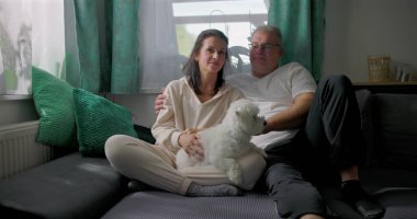 Küçük beyaz köpekle kanepede dinlenen, sakin ve samimi aile anlarının tadını çıkaran, doğal ışık ve yumuşak mobilyaları olan rahat oturma odası, sıcak atmosferi olan çift.