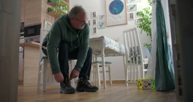 Babam, sıcak ahşap döşemeleri ve doğal ışıklandırması olan sıcak bir aile yemek odasında, emzik ile perdelerin yanında duran bir çocuğa bakarken botlarını bağlıyor.