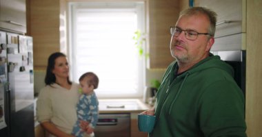 Annesi buzdolabını kollarında bebekle kontrol ederken elinde bardak tutan bir adam, tipik bir sabah rutini sırasında sıradan bir aile anı, sıcak ve besleyici ebeveynlik sahnesi.