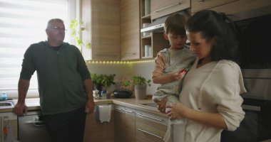 Anne çocuğunu kucağında bir sohbete tutarken, bir erkeğin arka plandan izlediği gibi rahat ve düzenli modern bir mutfakta.