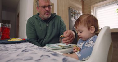Baba yemek sırasında bebek maşasında oturur, anne tabağın yanında durur, aile bağlarını yemek masasında kurar, çocuk yemek yer.