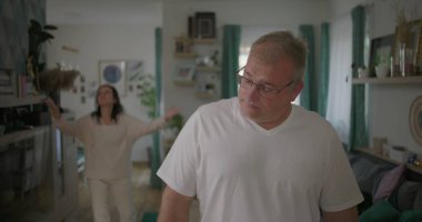 Erkek, arka plandaki kadınla tartışırken gözle görülür şekilde üzgün, oturma odasında ilişki gerilimi, duygusal ve dramatik anlaşmazlık anı