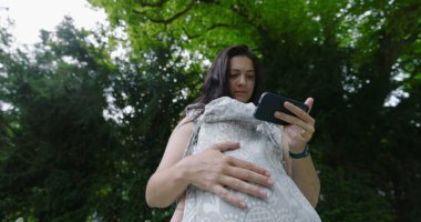 Parkta çoklu görev yapan bir kadın, akıllı telefonuna odaklanırken bebeğini bir taşıyıcıda tutuyor, ağaçlarla çevrili huzurlu bir açık hava ortamının tadını çıkarıyor.