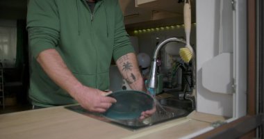 Bir adam mutfak lavabosundaki musluk suyunun altında mavi bir tabağı duruluyor, günlük ev işleri sırasında bulaşık takımlarını temizlemeye odaklanıyor.