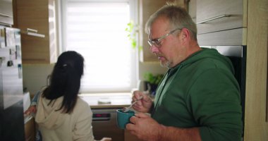 Anne bebeğini arka planda tutarken, adam mutfakta içki karıştırıyor. Aile sabah rutini sırasında birden fazla iş yapıyor. Günlük ev hayatı.