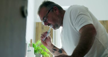 Adam lavaboda dişlerini fırçalıyor, hafifçe öne eğiliyor, temiz ve modern banyoda sabah rutininin bir parçası olarak kendine bakım ve ağız temizliğine odaklanıyor.