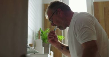 Banyo lavabosunda dişlerini fırçalayan adam, yoğun ifadeli yan profil görüntüsü, parlak ve temiz ev ortamında sabah diş bakımı rutini