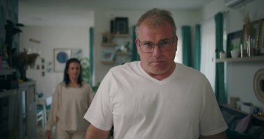 Erkek, tartışma sırasında yoğun bir ifadeyle kameraya bakıyor, kadın oturma odasında, gergin ve duygusal bir ilişki çatışması ve hüsran anı yaşıyor.