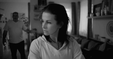 Kadın hararetli tartışmalar sırasında erkeklerden yüz çeviriyor, mesafeli ve üzgün görünüyor, erkek ise arka planda hayal kırıklığıyla işaret ediyor, duygusal gerilimi ve çatışmayı yakalıyor.