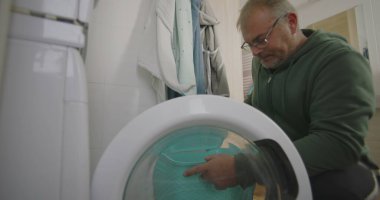 Çamaşır makinesinin önünde diz çökmüş bir adam, temiz havluları ve parlak döşeli duvarları olan modern bir banyoda çamaşırları dikkatlice yüklüyor.