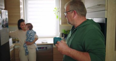 Anne bebeğini arka planda tutarken, adam mutfakta içki karıştırıyor. Aile sabah rutini sırasında birden fazla iş yapıyor. Günlük ev hayatı.