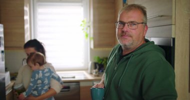 Annesi buzdolabını kollarında bebekle kontrol ederken elinde bardak tutan bir adam, tipik bir sabah rutini sırasında sıradan bir aile anı, sıcak ve besleyici ebeveynlik sahnesi.