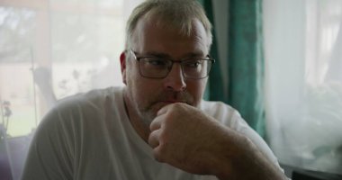 Adamın biri, çenesini elinde dinlendirirken, sakin ve rahat bir evde yumuşak yeşil perdeleri olan bir pencerenin yanında otururken düşünceli bir şekilde bakıyor.