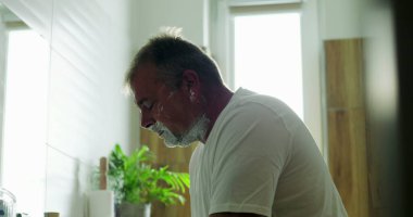 Jiletle tıraş olan, çene açısını hassasiyete göre ayarlayan, yüzdeki köpüğü tıraş eden düşünceli ifade, banyo arka planı, sabah bakım rutini.