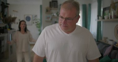 Oturma odasında hararetli tartışmalar yaşayan bir çift, görünürde üzgün duran bir adam, arka planda duygusal hareketler yapan bir kadın, ilişki çatışması sırasında gergin bir an.