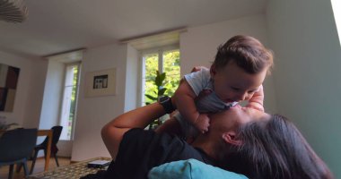 Anne, anne ve çocuk arasındaki bağ ve sevgi dolu doğal ışıkla dolu parlak bir oturma odasında oynarken, gülümserken ve gülerken bebeği havaya kaldırıyor.