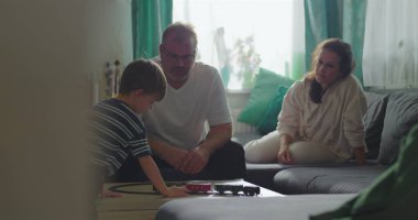 Baba kahve masasında oyuncak trenle oynayan çocuğu izliyor, anne arka planda küçük beyaz bir köpek tutuyor, aile bağı ve sakin bir oturma odasında oyun zamanı.