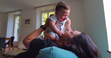 Anne, anne ve çocuk arasındaki bağ ve sevgi dolu doğal ışıkla dolu parlak bir oturma odasında oynarken, gülümserken ve gülerken bebeği havaya kaldırıyor.