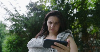 Anne endişeli bir ifadeyle, bebeğini bir taşıyıcıda tutarken dikkatle parktaki akıllı telefonuna odaklanıyor, modern endişelerin ve ebeveynliğin karışımını vurguluyor.