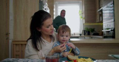 Anne gülümserken, bebek kahvaltıda sarı dolmalık biber ararken sıcak ve besleyici atmosferli sıcak bir mutfakta, aile yemeği eşliğinde kaynaşıyor.