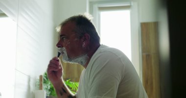 Jiletle tıraş olan, çene açısını hassasiyete göre ayarlayan, yüzdeki köpüğü tıraş eden düşünceli ifade, banyo arka planı, sabah bakım rutini.