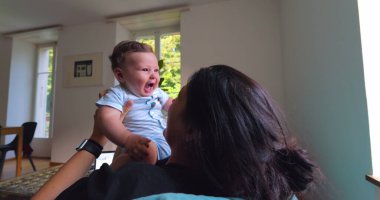 Anne, anne ve çocuk arasındaki bağ ve sevgi dolu doğal ışıkla dolu parlak bir oturma odasında oynarken, gülümserken ve gülerken bebeği havaya kaldırıyor..