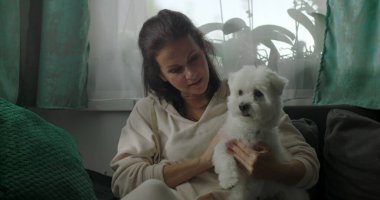 Kanepede oturan bir kadın kucağında küçük beyaz bir köpek, nazik bir ifadeyle aşağıya bakıyor, evde sakin bir bağ kurma anı, sakin bir evde yumuşak bir arkadaşlık.