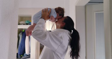 Anne sevinçle bebeğini havaya kaldırıyor, aydınlık bir koridorda sevgi dolu bir anı paylaşıyor. neşeli etkileşimler ve gülümsemeler anne ve çocuk arasındaki güçlü bağı ve mutluluğu gösterir.