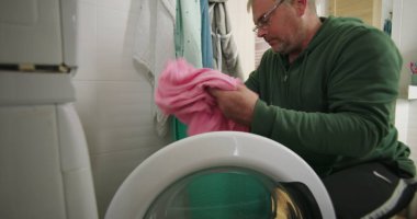 Çamaşır makinesine çamaşır sepeti ve temiz ve modern banyo ortamında havluları koymaya hazırlanırken gülümseyen bir adam.