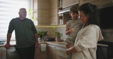 Anne çocuğunu kucağında bir sohbete tutarken, bir erkeğin arka plandan izlediği gibi rahat ve düzenli modern bir mutfakta.