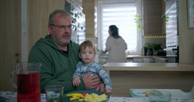 Babam yemek masasında renkli yemek ve içeceklerle bebeği tutarken annem de mutfakta, sıcak aile ortamında sıcak aile ortamını hazırlıyor.