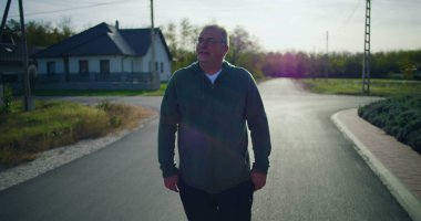 Huzurlu bir banliyö sokağında yan bakan orta yaşlı bir adam, gözlük takıyor ve sakin bir mahallede yansıtıcı ve düşünceli bir tavır sergiliyor.