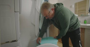 Çamaşır makinesinin önünde çömelmiş bir adam, çamaşırları küçük bir banyodaki çamaşır makinesine yüklüyor, ev işlerini odaklanarak ve verimlilikle yapıyor.