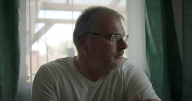 Pencere kenarında oturan bir adam, derin düşüncelere dalmış, elleri çapraz, sakin atmosfer kapalı, pencere ve perdeler yansıtıcı bir ruh halini çerçeveliyor.