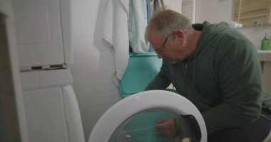 Çamaşır makinesinin önünde çömelmiş bir adam, çamaşırları küçük bir banyodaki çamaşır makinesine yüklüyor, ev işlerini odaklanarak ve verimlilikle yapıyor.